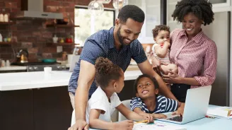 A family of five is in the kitchen, helping two of their children with their homework.