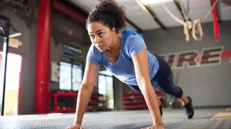 A young African American woman with a blue shirt performs push-ups at an open spaced gym.