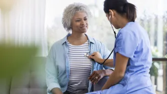 A female doctor is seated, listening to a senior woman's heart with a stethoscope.