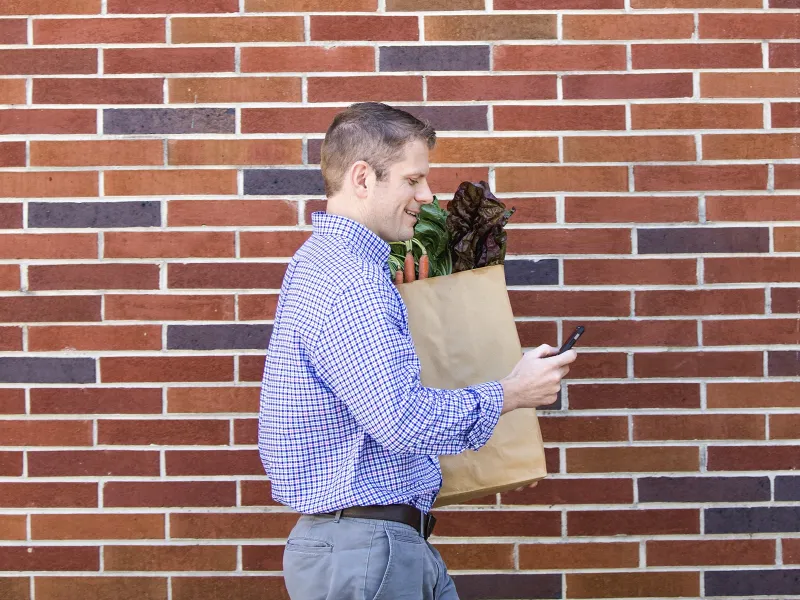 A young Caucasian man checks his cell phone while carrying a bag of fresh produce.