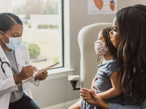 Mother and Child Visit Their Pediatrician in an Exam Room in Front of a Large Window While Wearing Face Masks.