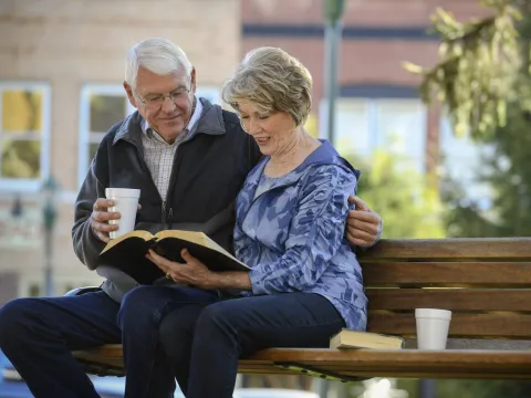 Elderly couple sitting on a park bench reading the bible. 